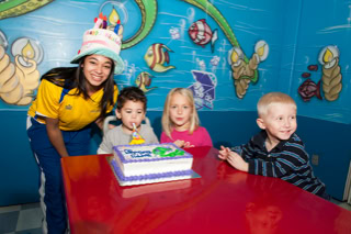 A group of children sitting at a red table with a birthday cake, accompanied by an adult wearing a festive hat. The background features colorful, aquatic-themed wall decorations.