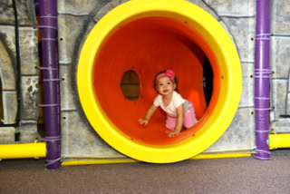 A child wearing a pink outfit and headband is crawling inside a large, yellow and orange play tunnel.