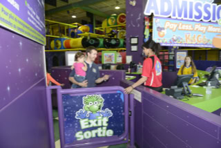 A man holding a child is standing at a counter in an indoor play area with colorful decorations. A staff member in a red shirt is behind the counter, and a sign with "Exit Sortie" is visible.
