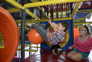 Child playing on an orange ball in an indoor playground, with another child watching nearby.